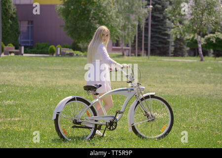 Très jolie jeune femme en robe blanche avec vélo sur sunny vert prairie horizons revient sur camera Banque D'Images