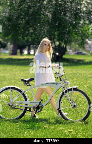 Très jolie jeune femme en robe blanche avec vélo sur sunny vert prairie horizons examine l'appareil photo Banque D'Images