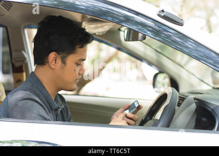 Young attractive asian male en utilisant un téléphone portable au volant de sa voiture à sunny day Banque D'Images