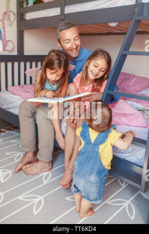 Famille de quatre membres des gens assis sur lit dans la chambre lecture livre. Père et filles les filles à passer du temps ensemble. Parler parent commun Banque D'Images