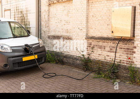 Voiture de couleur noir blanc debout à côté d'un mur est électriquement chargés par l'intermédiaire d'un câble connecté à un point de connexion accroché sur un mur de pierre Banque D'Images