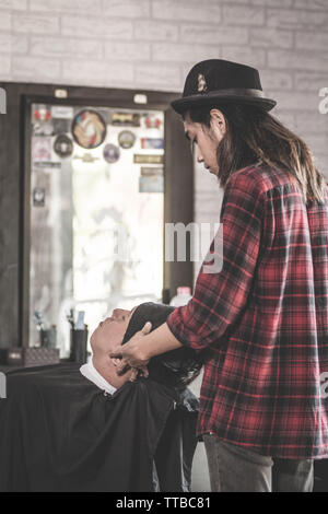 Vue arrière du salon de coiffure homme avec de longs cheveux massage aux clients de faire avec des serviettes qui vous détendre assis au fauteuil de barbier Banque D'Images