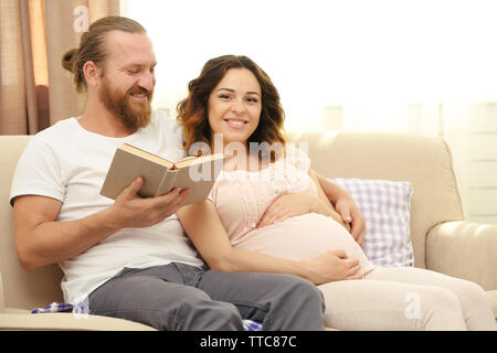 Bel homme lit livre à sa charmante femme enceinte sur le canapé dans la chambre Banque D'Images