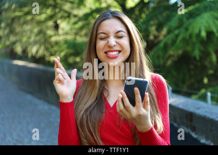 Fille d'espoir crossing fingers holding un smartphone dans l'attente de nouvelles à l'extérieur. Jeune femme avec des doigts de passage et smart phone qui souhaitent le meilleur outsid Banque D'Images