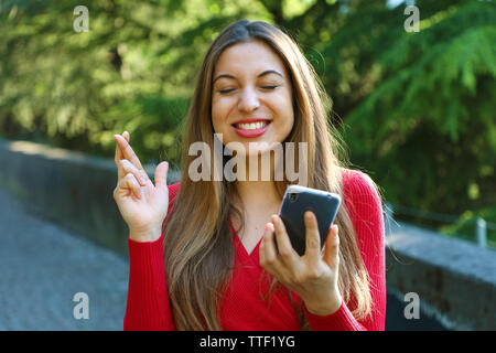 Fille d'espoir crossing fingers holding un smartphone dans l'attente de nouvelles à l'extérieur. Jeune femme avec des doigts de passage et smart phone qui souhaitent le meilleur outsid Banque D'Images