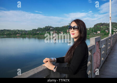 Portrait de femme portant des lunettes de soleil alors qu'il se trouvait sur la passerelle sur la rivière contre le ciel bleu Banque D'Images