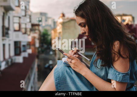 Vue latérale du young woman using smart phone en position assise contre les bâtiments en ville Banque D'Images