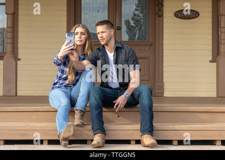 Jeune couple d'usure de l'ouest sur le porche de la maison en tenant vos autoportraits Banque D'Images