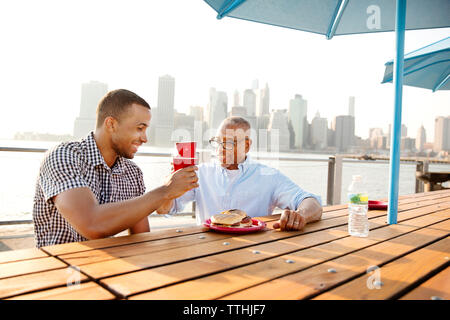 Heureux père et fils toasting drinks assis à table par l'East River en ville Banque D'Images