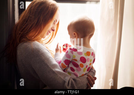 Femme regardant bébé fille jouant avec ornement de noël tout en se tenant par la fenêtre à la maison Banque D'Images