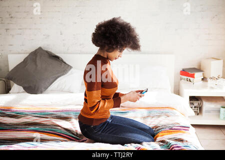 Vue latérale de la femme à l'aide de smart phone tout en s'agenouillant sur le lit chez lui Banque D'Images