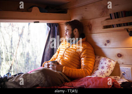 Smiling man with mug looking away while sitting on bed in camper van Banque D'Images