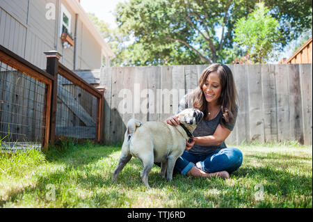 Femme jouant avec le pug while sitting on grassy field in backyard Banque D'Images