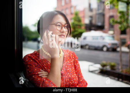 Happy Woman talking on smart phone while standing by window Banque D'Images
