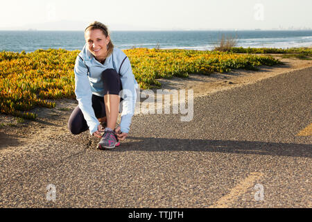 Portrait de femme tout en lacet attachant accroupi sur route contre la mer Banque D'Images