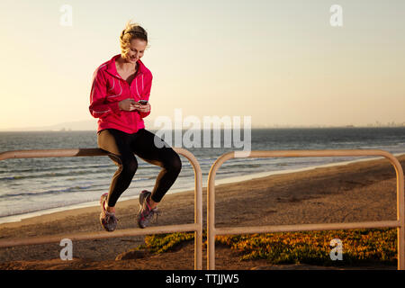 Femme à l'aide de smart phone while sitting on railing at beach against clear sky Banque D'Images