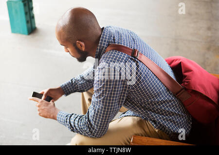 Portrait de l'homme à l'aide de smart phone while sitting on bench à railroad station Banque D'Images