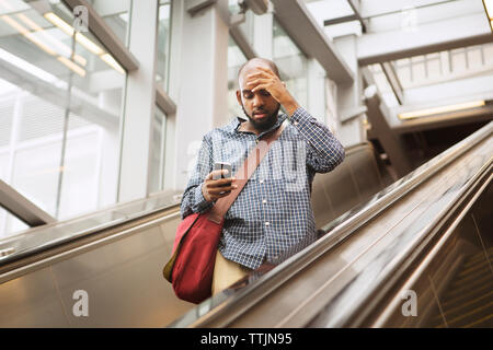 L'homme avec la tête dans la main à l'aide de smart phone while standing on escalator à railroad station Banque D'Images