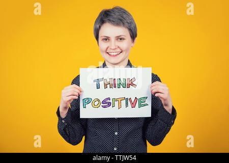 Extraverti optimiste hipster Jeune femme tenant une feuille de papier blanc avec le texte du message pensez positif plus isolé sur fond jaune. Mo colorés Banque D'Images