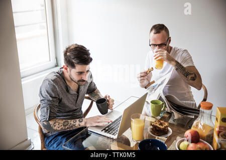 Les hommes gais à l'aide d'ordinateur portable et lisant le journal pendant le petit-déjeuner à la maison Banque D'Images