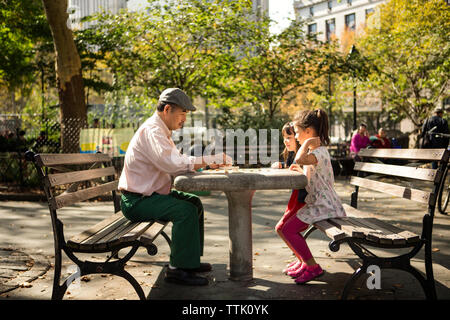 Vue latérale du grand-père et petites-filles joue une partie tout en étant assis sur un banc dans le parc Banque D'Images