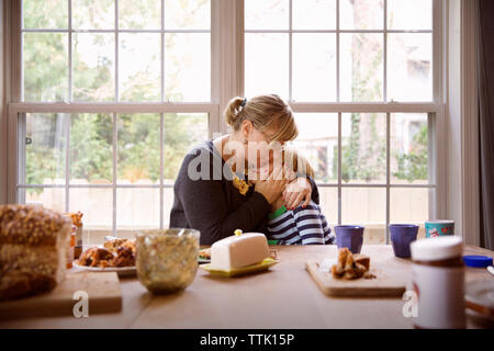 Mère embrassant fils assis à table des aliments contre la fenêtre dans salle de séjour Banque D'Images
