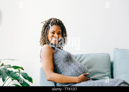 Portrait of smiling pregnant woman sitting on sofa par wall à la maison Banque D'Images