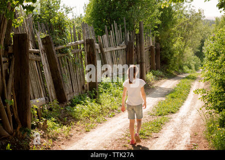 Vue arrière de fille qui marche sur la route de terre par jour ensoleillé au cours de clôture en bois Banque D'Images
