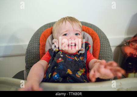 Portrait de bébé garçon malpropre de pleurer alors qu'il était assis sur la chaise haute contre le mur à la maison Banque D'Images