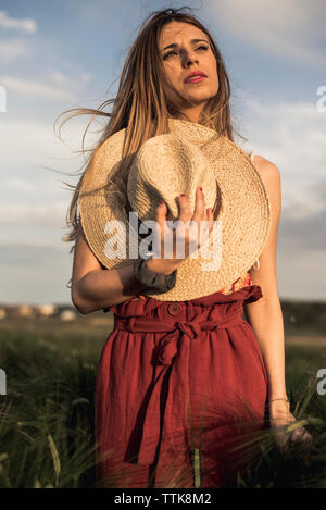 Low angle view of thoughtful woman holding hat while standing on grassy field against sky Banque D'Images