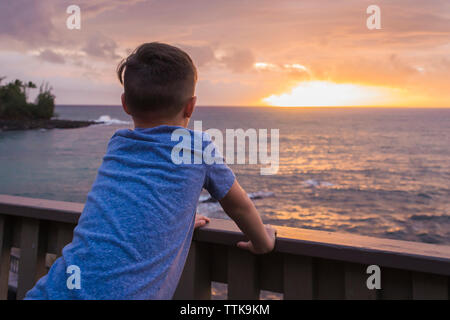 Vue arrière du garçon à la recherche en mer alors qu'il se tenait en main courante des pier pendant le coucher du soleil Banque D'Images