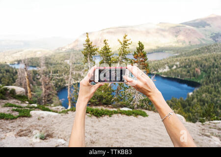 Mains coupées d'amies en tenant avec selfies sur smart phone contre montagne ciel en forêt Banque D'Images