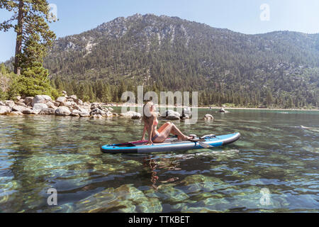 Vue de côté de woman in bikini paddleboarding sur le lac contre Mountain au cours de journée ensoleillée Banque D'Images