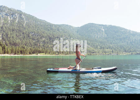 Vue de côté de woman in bikini paddleboarding sur le lac contre ciel clair au cours de journée ensoleillée Banque D'Images