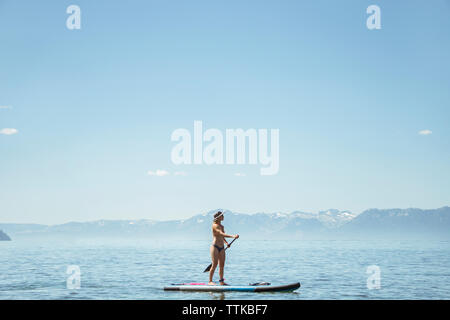 Vue de côté de woman in bikini paddleboarding sur le lac contre le ciel bleu au cours de journée ensoleillée Banque D'Images