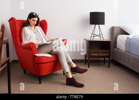 Businesswoman reading magazine alors qu'il était assis sur un fauteuil rouge dans la chambre d'hôtel Banque D'Images