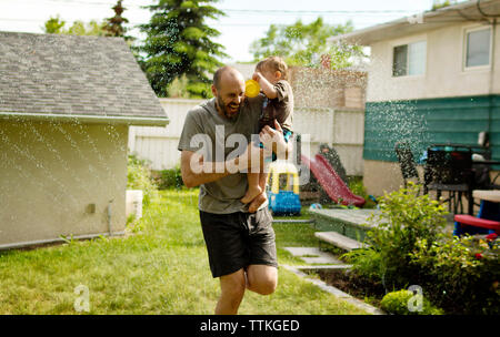 Cheerful father carrying son lors de la lecture en arrière-cour au sprinkleur Banque D'Images