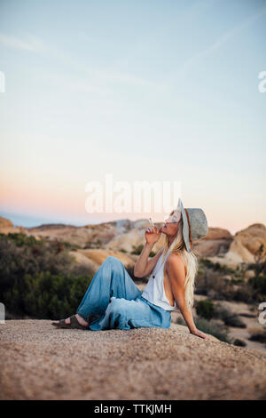 Femme fumant alors qu'il était assis sur la roche à Joshua Tree National Park against sky Banque D'Images