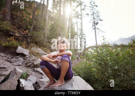 Happy girl sitting on rock against trees in forest Banque D'Images