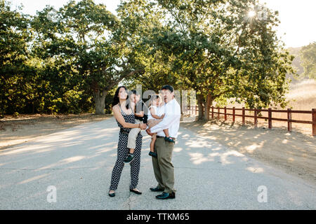 Portrait of happy parents transportant des enfants en étant debout sur la route contre des arbres en parc Banque D'Images