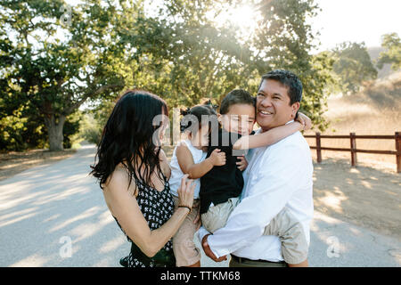 Heureux parents multi-ethnique transportant les enfants mignons alors que l'article sur la route contre des arbres en parc Banque D'Images
