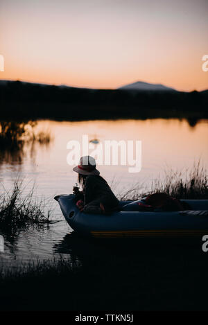 Fille assise en radeau sur le lac contre ciel clair pendant le coucher du soleil Banque D'Images