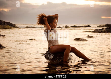 Teenage girl sitting on rock avec la main dans la mer au milieu de cheveux Banque D'Images