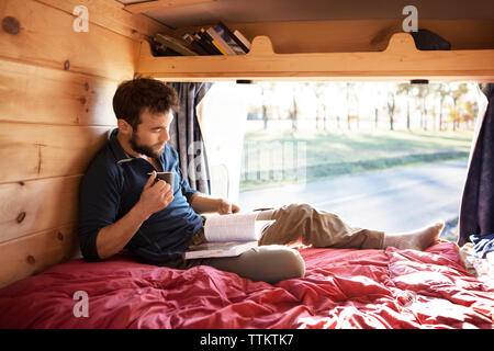 Homme avec mug reading book while sitting on bed in camper van Banque D'Images