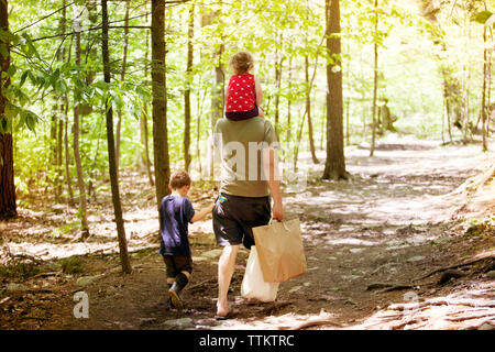 Vue arrière du père holding sacs avec son fils et sa fille marche sur chemin de terre, au milieu des forêts Banque D'Images