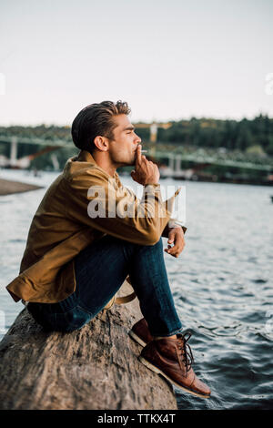 Vue latérale du man smoking while sitting on log par river contre ciel clair Banque D'Images