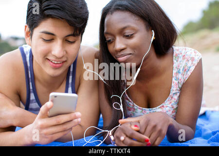 Jeune couple à l'écoute de la musique via smart phone while lying on blanket at beach Banque D'Images