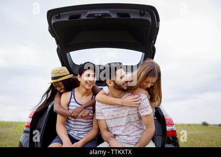 Cheerful friends sitting in car trunk sur terrain contre ciel clair Banque D'Images