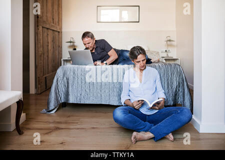 Senior couple using laptop and reading book in bedroom Banque D'Images