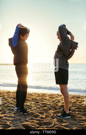 Toute la longueur de père et fils d'armes d'étirement en position debout face à face à l'encontre de la plage pendant le coucher du soleil Ciel clair Banque D'Images